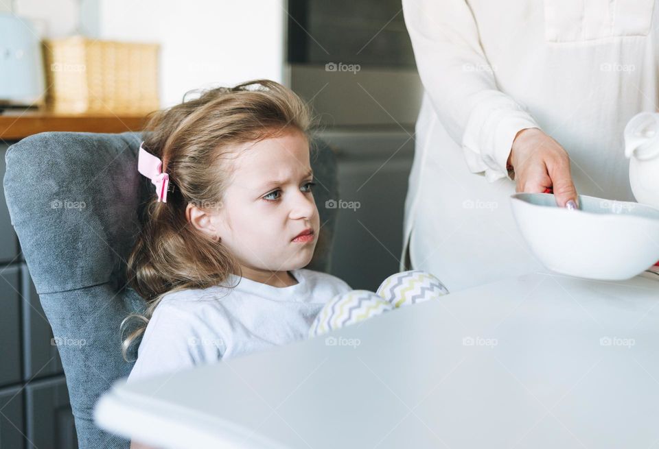 Unhappy little child beautiful girl with long hair in home clothes with yogurt sitting at dinning table in kitchen at home. Child does not want to eat