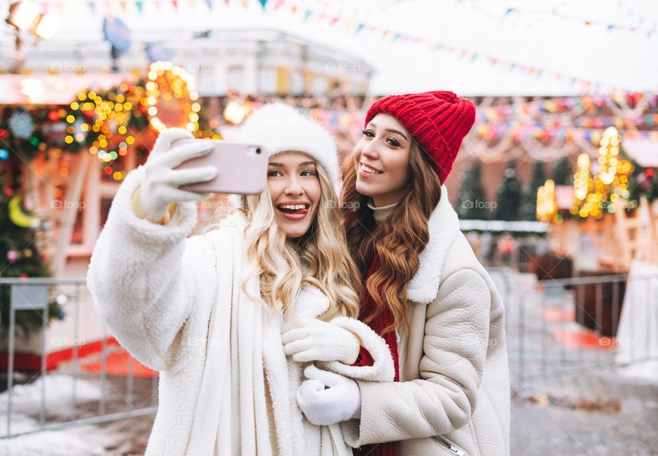 Young happy women friends with curly hair in red having fun taking selfie on mobile phone in winter street decorated with lights