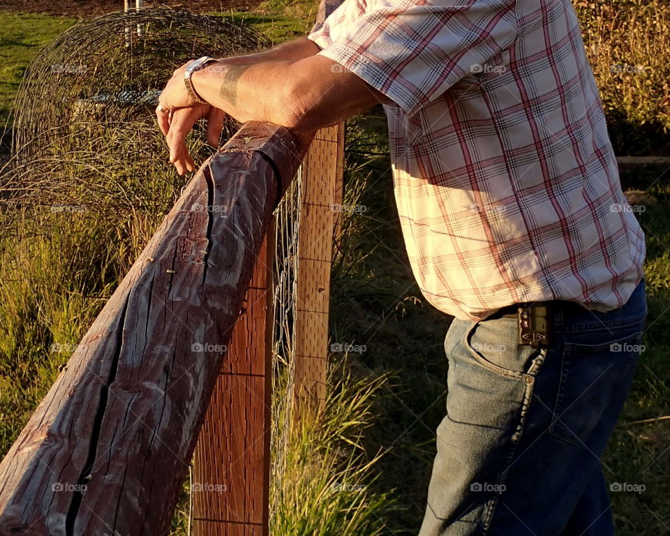 A man leans against a fence on his farm in Central Oregon watching the sun set across his field. 