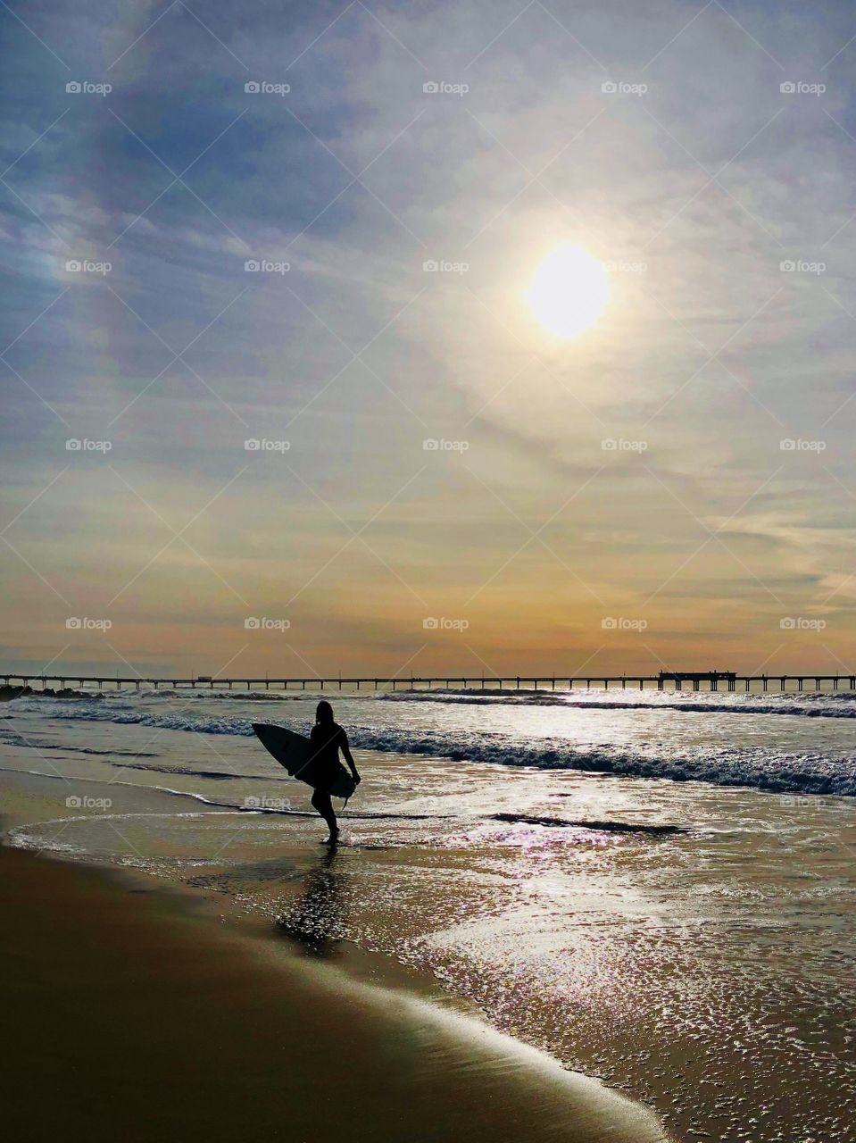 The silhouette of a surfer emerges from the water, bright lit sky behind them. Light bounces off the ocean while the sun begins to set.