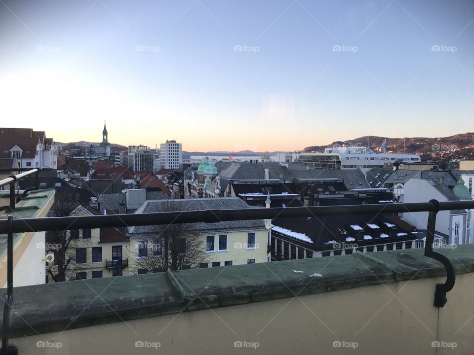Sunset in Bergen, Norway. The sky is clear, and the  orange sunset fades into the blue sky. No clouds in Bergen. The roofs of the city shows way to the sunset. One of the church towers in Bergen shows clearly in the background.