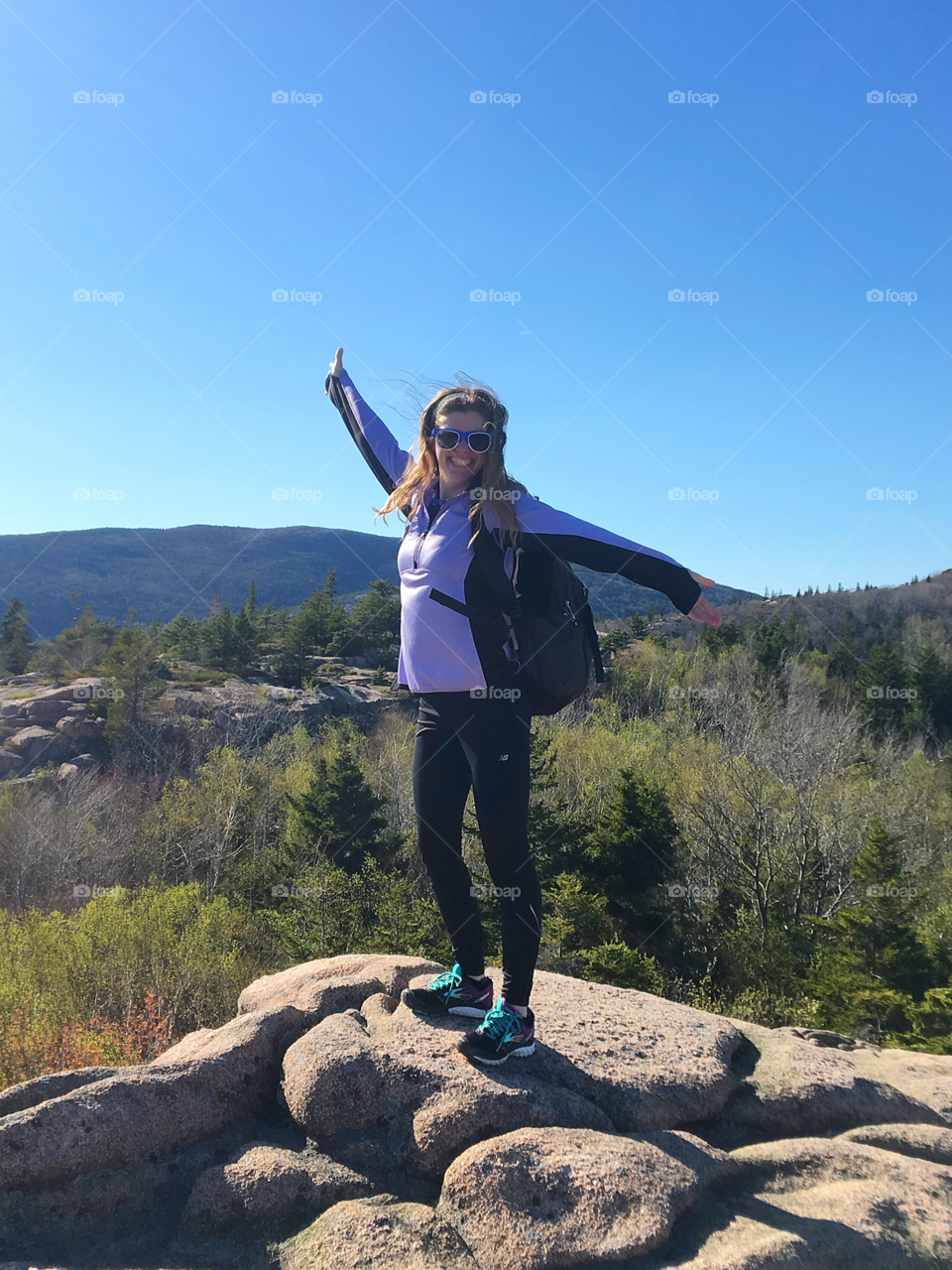 A woman stands triumphantly at the summit of a mountain hiking trail. The mountain woods below show the first signs of spring