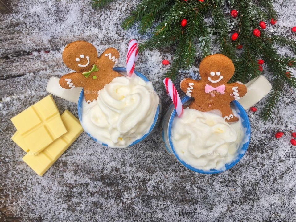 Two mugs of white chocolate with whipped cream, gingerbread man and candy canes on a snowy table with pieces of white chocolate and evergreen branches beside