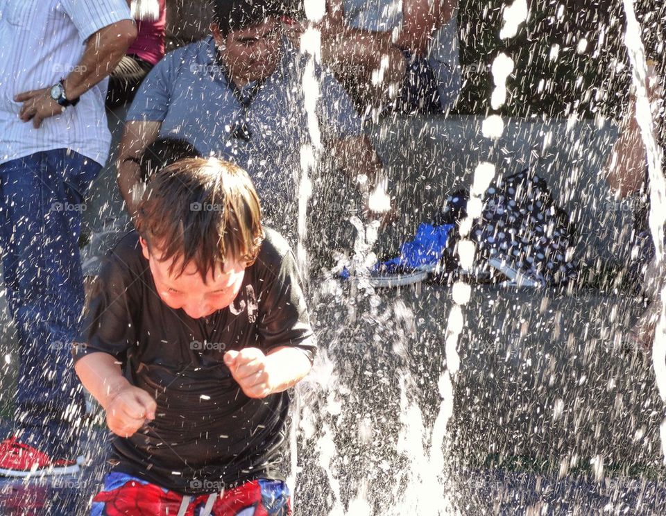 Boy Splashing In Water Fountain. Cooling Off In The Summer