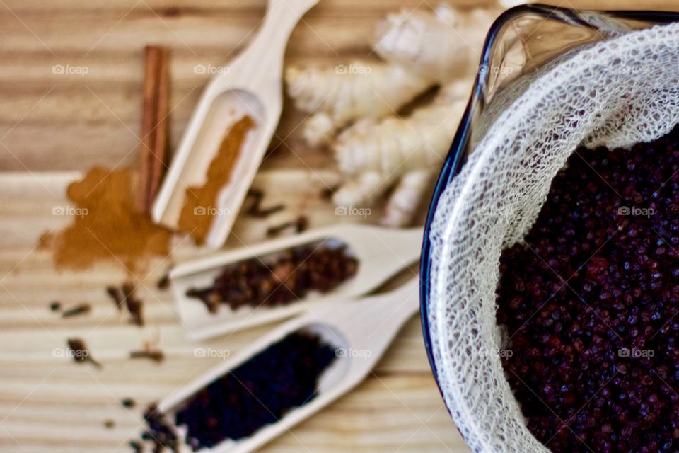 Overhead view of elderberries being strained for elderberry syrup in a colander lined with cheesecloth over a large glass measuring cup, blurred background including whole and ground spices in wooden scoops on a wooden surface
