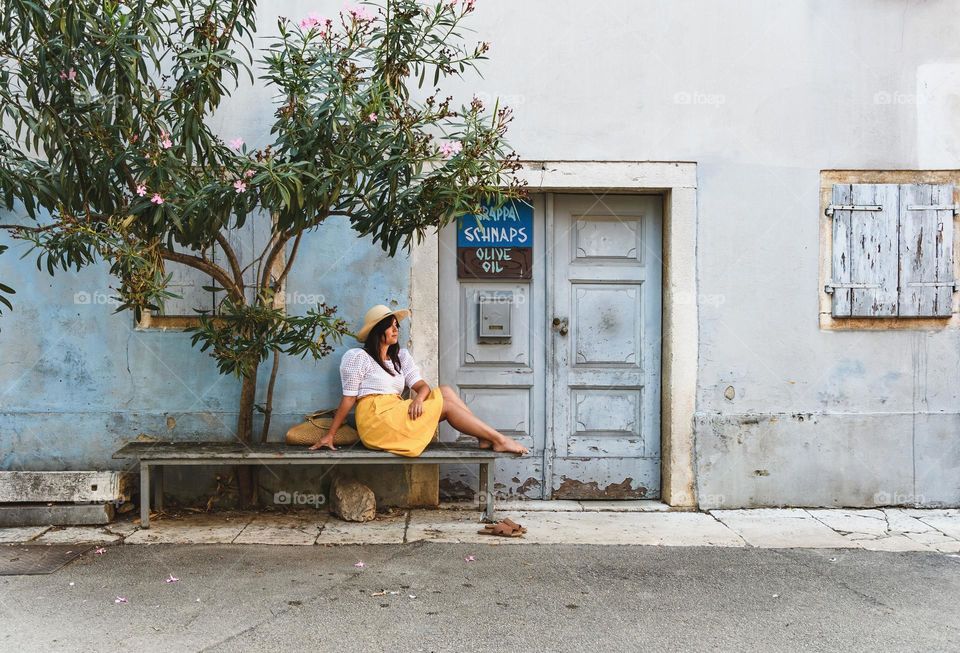 Young woman sitting on bench under oleander tree in font of old house