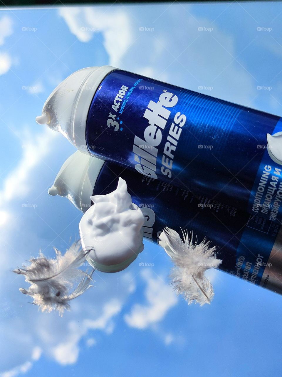 Men's cosmetics. On a mirrored surface lies a blue bottle with a white inscription: "Gillette series", two bird feathers and white fluffy foam. The mirror surface reflects objects on the mirror surface and the blue sky with white clouds