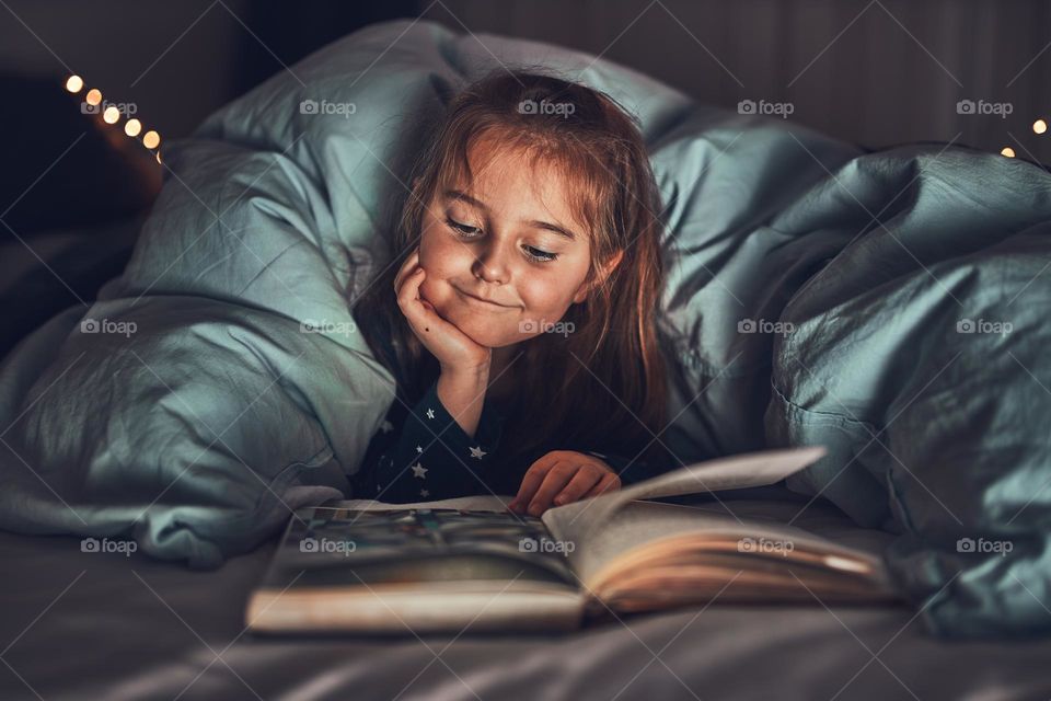 Little girl reading book and watching pictures in bed before going to sleep. Reading stories before sleep. Bedtime stories for child