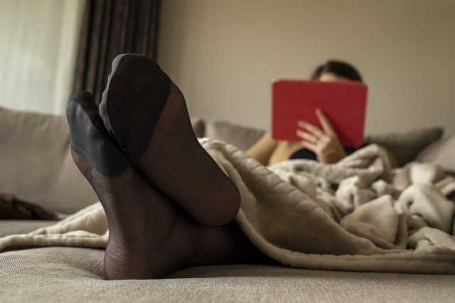 A portrait of the black pantyhose feet of a girl lying under a blanket on a couch looking at her tablet.