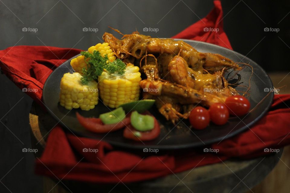 Asian food balado fried prawns with corn chunks, lemon slices and tomatoes served on black plate with red cloth bottom and wooden coaster. Asian style street seafood.