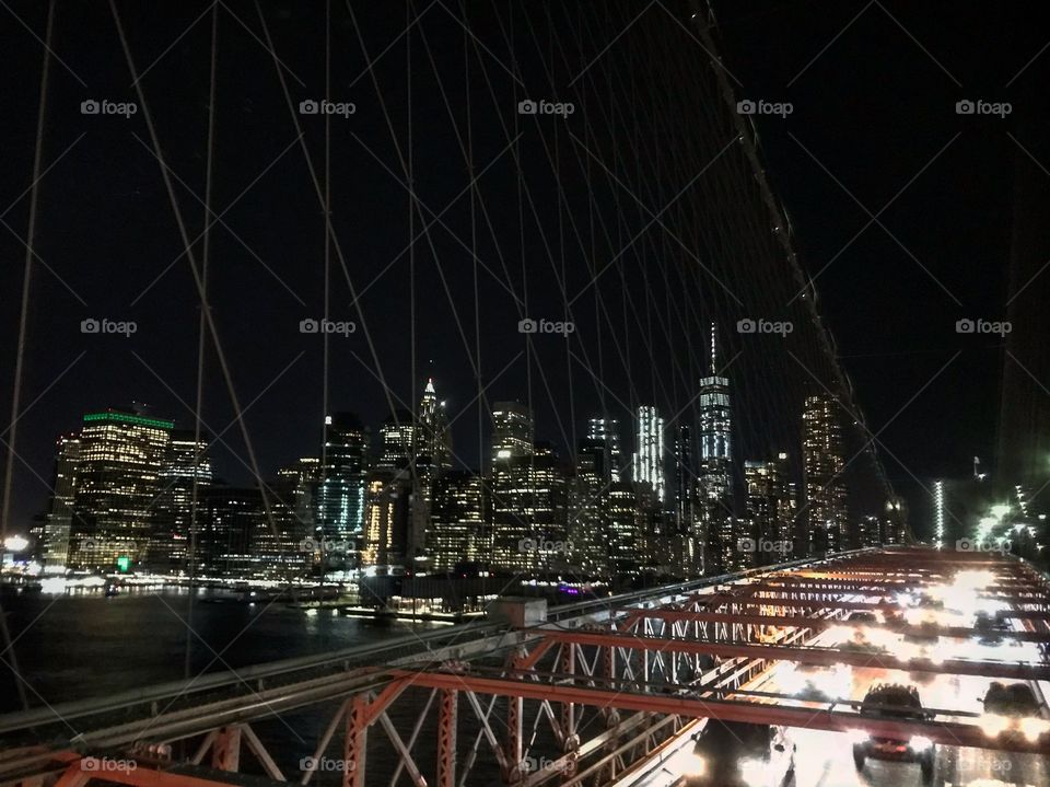 Nighttime photo of the New York City skyline taken from the Brooklyn Bridge. Some of the cars on the bridge and bridge cables are also seen. 
