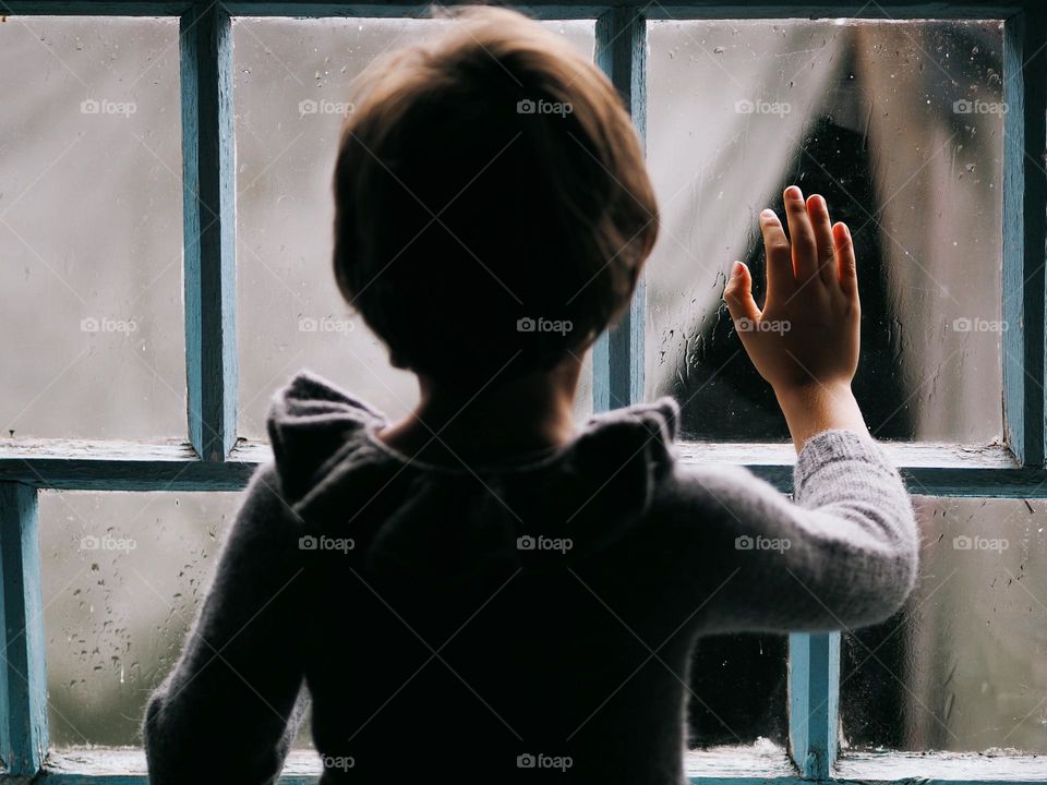 Little boy looking out window at home on a rainy day, wearing grey knitted sweater. Child stuck inside on a rainy day, touching the window and looking out the window at the rain falling on the street, back view.