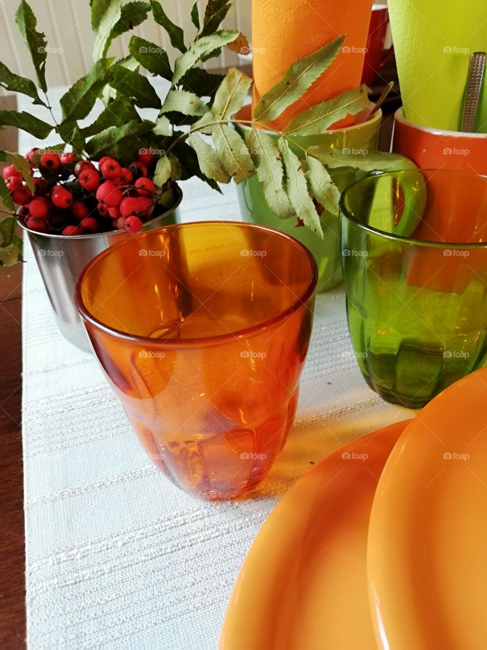 Couple of green and orange glasses and ceramic mugs, two orange plates on a light blue table cloth, which is embossed. In the cups napkins and teaspoons, a branch of rowan berries in a silver color tin.