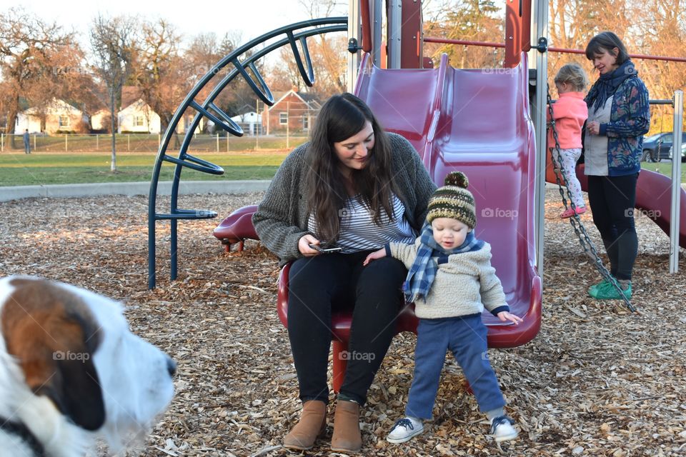 Cute toddler boy playing on playground slide with his mother while their loyal pet dog watches 