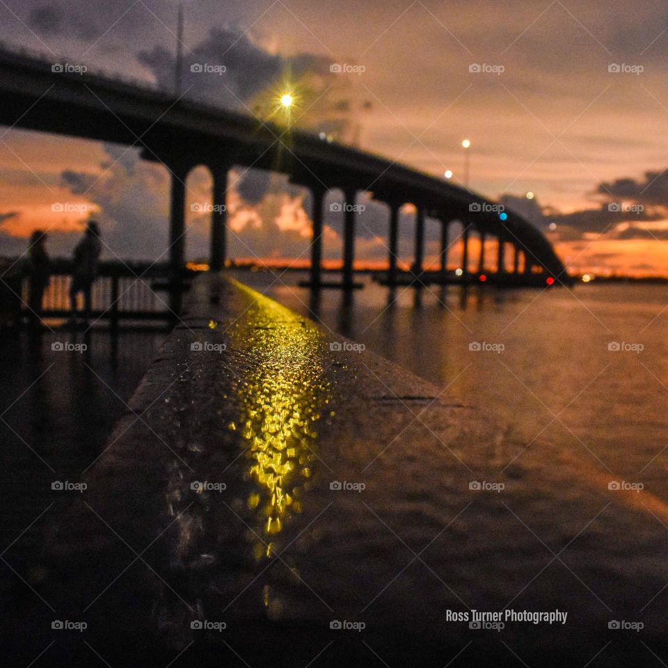 A streak of light is shown in focus in the foreground as it is reflected on a wooden railing on the fishing pier at sunset. The bridge is seen in the background as a silhouette against the orange and purple sky, with lights shining from the poles.