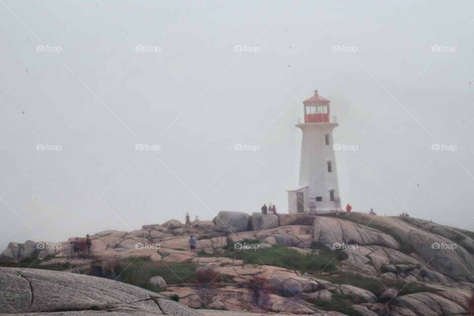 Long exposure photograph of tourists walking about on the rocks surrounding Peggy's Cove Lighthouse on a foggy afternoon.