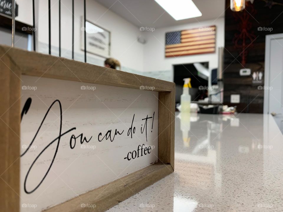 « You can do it! -coffee » sign in the foreground focus at a coffee shop interior small business unique brand with coffee maker workers on the move with happiness seeing the coffee cups holder with the American flag on the background wall.