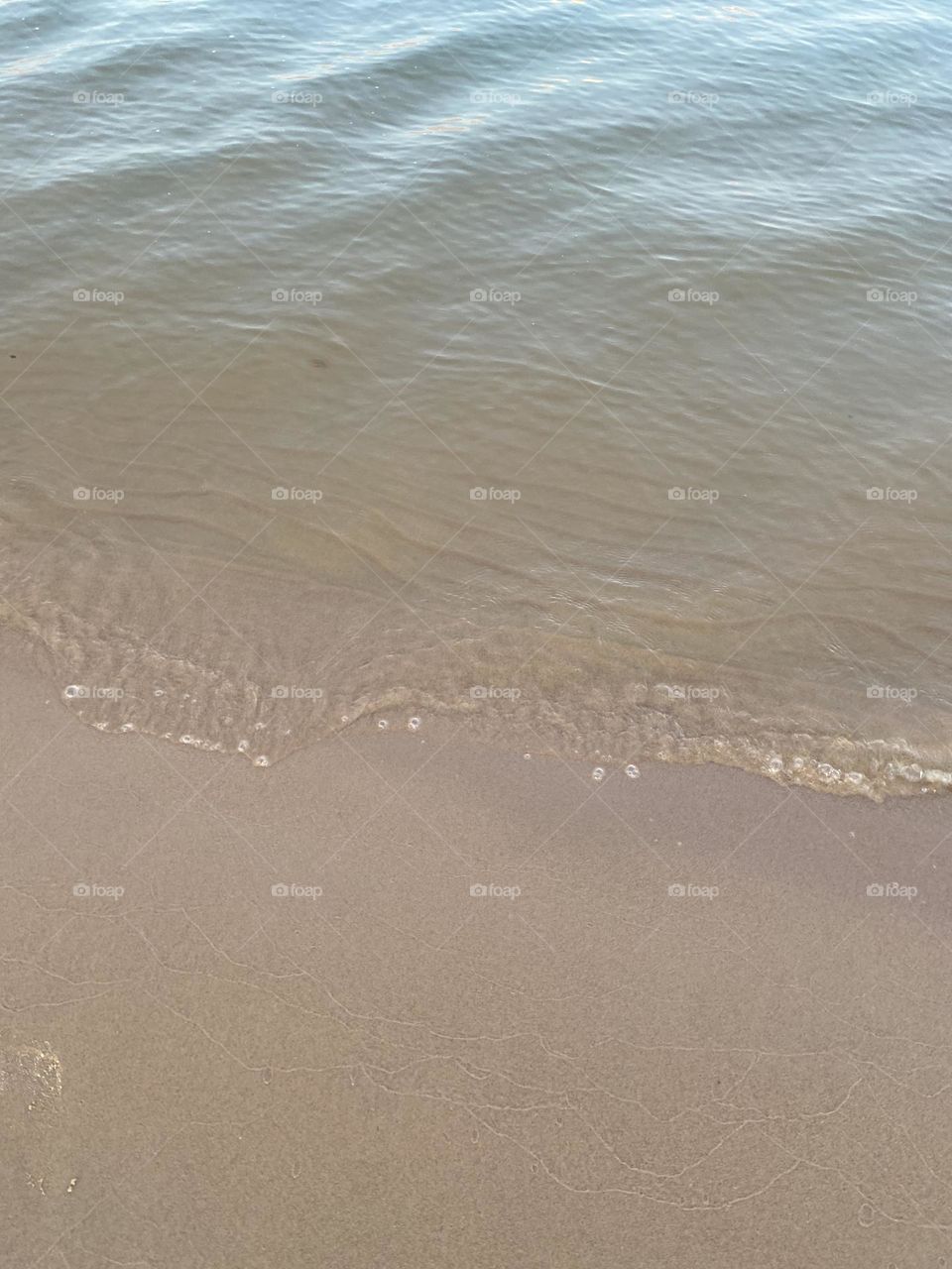 This image captures a close-up view of gentle waves lapping against a sandy beach. The water appears calm, and the shoreline is smooth with small bubbles forming at the edge where the water meets the sand, a peaceful and serene beach scene.