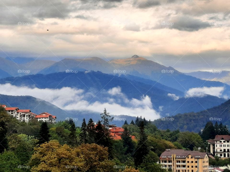 A calm landscape in a mountainous place, a view of low clouds, colorful roofs of houses and the autumn color of the foliage of trees