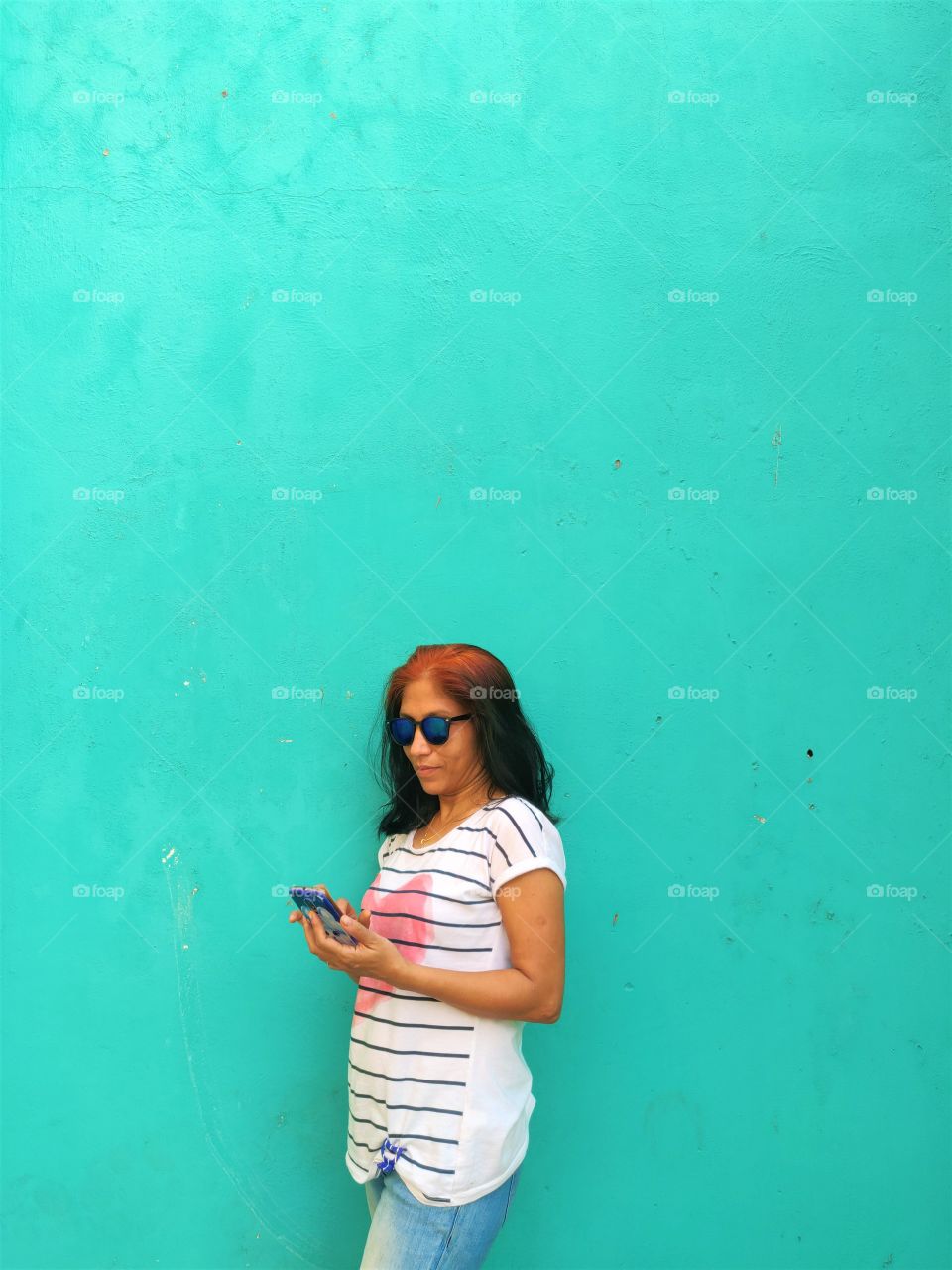 woman wearing white top standing in front of a green color wall and using her smartphone