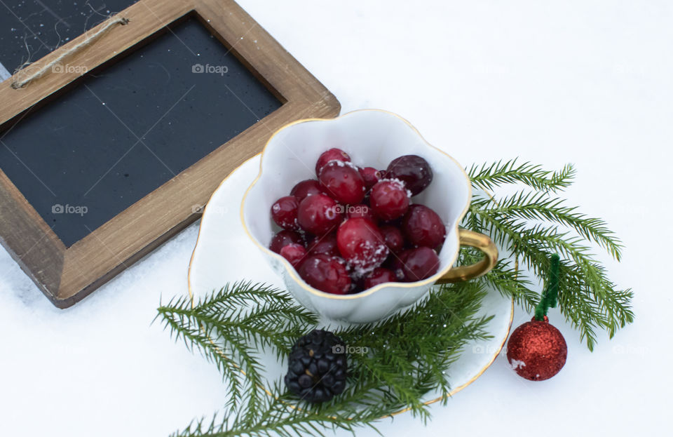 Cranberries in porcelaine vintage teacup in snow with evergreen decoration and blackboard with wood frame with room for copy or holiday greeting
