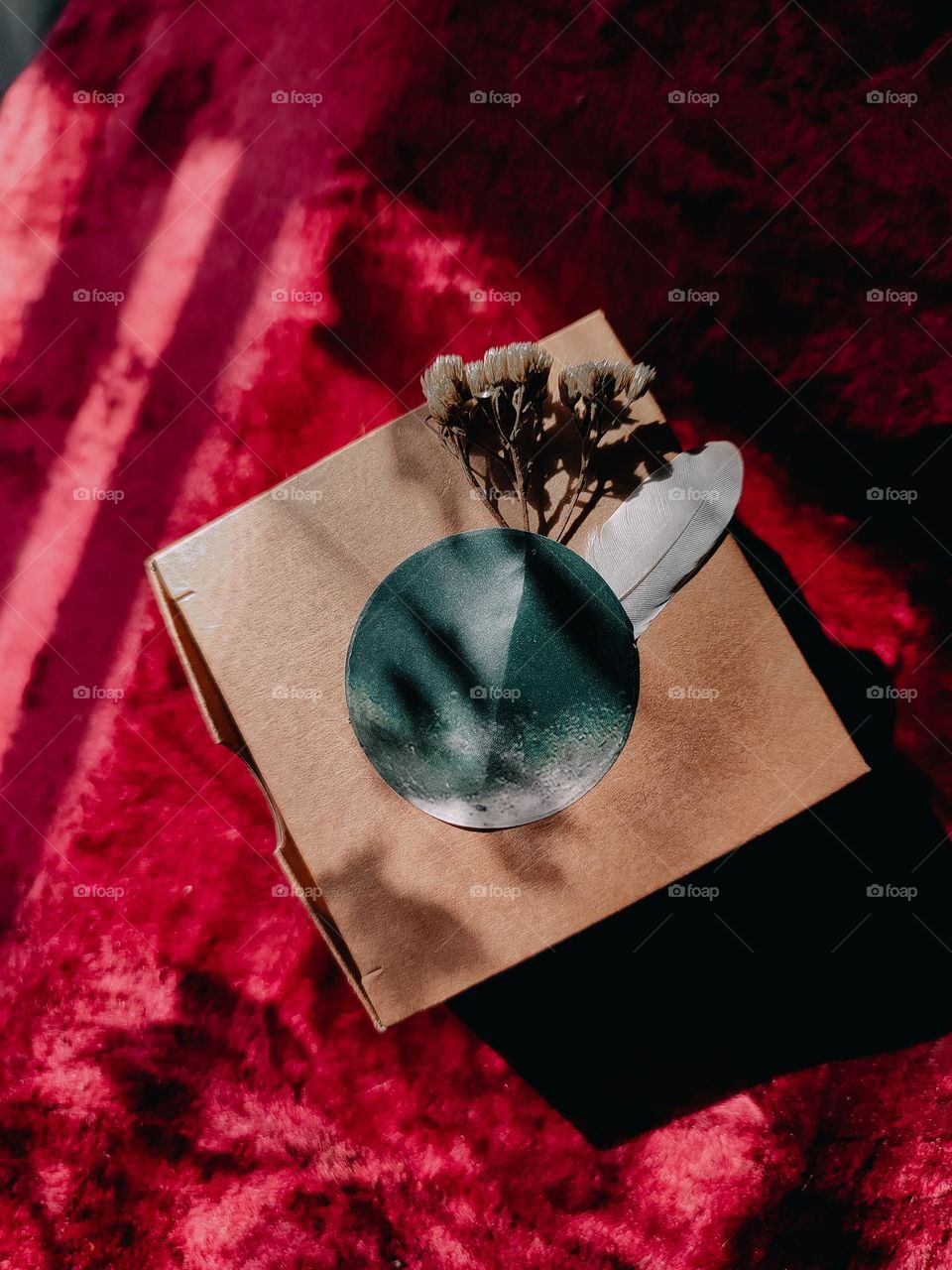 Brown craft paper box for jewelry with moon sticker, dry flowers and feather decor on the red velvet background. Sun rays shine from the window