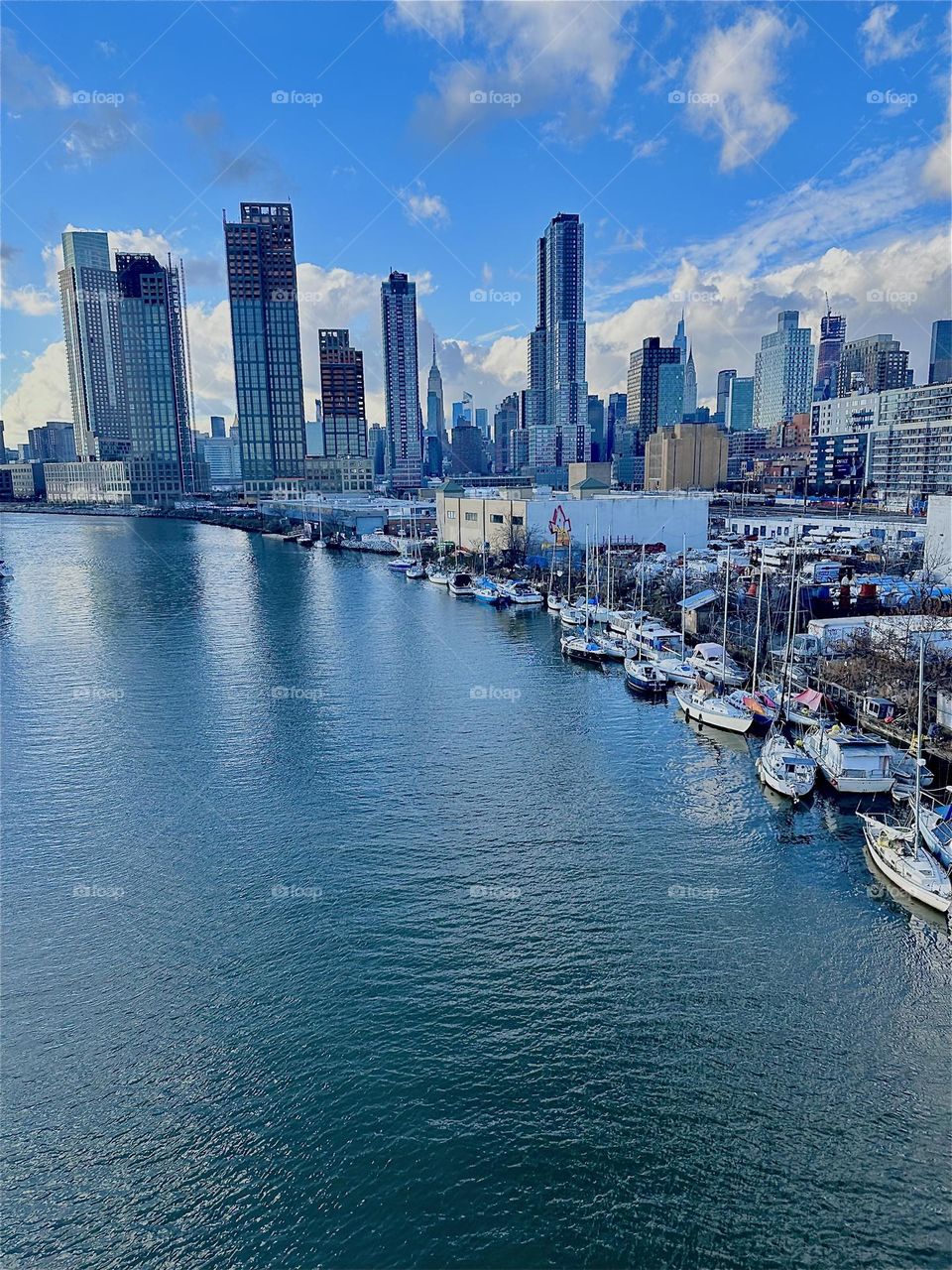 This is the view of “Newtown Creek” from the “Pulaski Bridge” that connects LIC, Queens to “Greenpoint”, Bklyn. A variety of boats is tied to the shore here. In the distance we see “Manhattan” incl. the “Empire State Bldg”. 2023. Hypnotic Productions