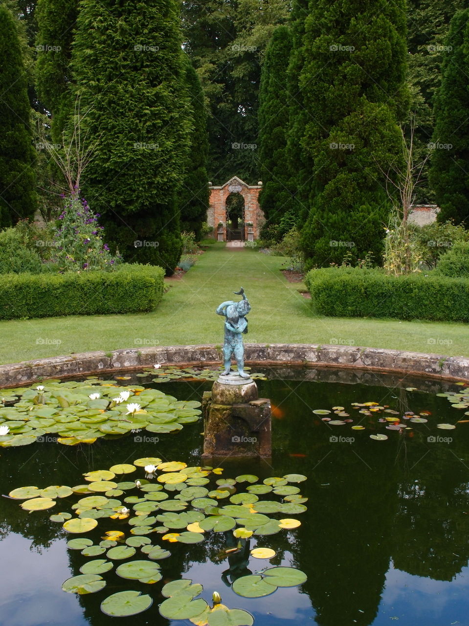 Reflections and floating Lilly Pads in a fountain in lushly landscaped gardens with shaped bushes, manicured lawns, a statue, and a stone arch on a summer day in England. 