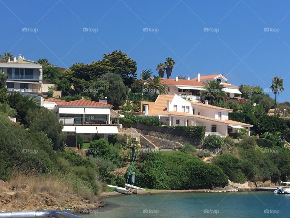 Some of the villas used by the wealthy residents of Mahon, as well as some tourists. A simple sailing boat can be seen resting on the beach by the water’s edge.