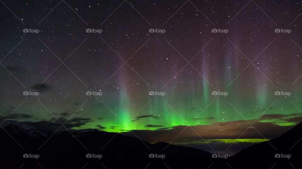 The northern lights shine high over the clouds in the night sky, with green and purple colour and many visible stars.