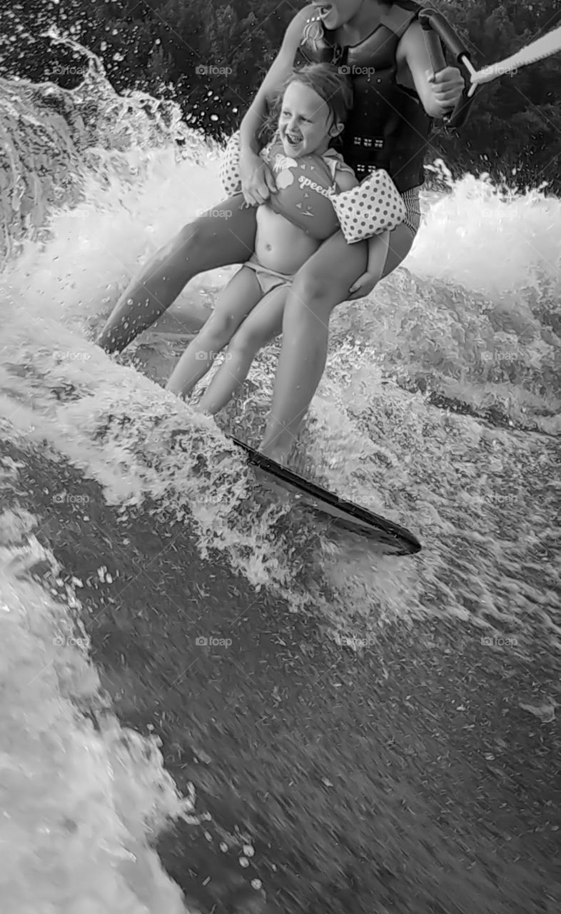 A little girl holds on tight to a woman while they hit a wave while surfing behind a boat on lake.
