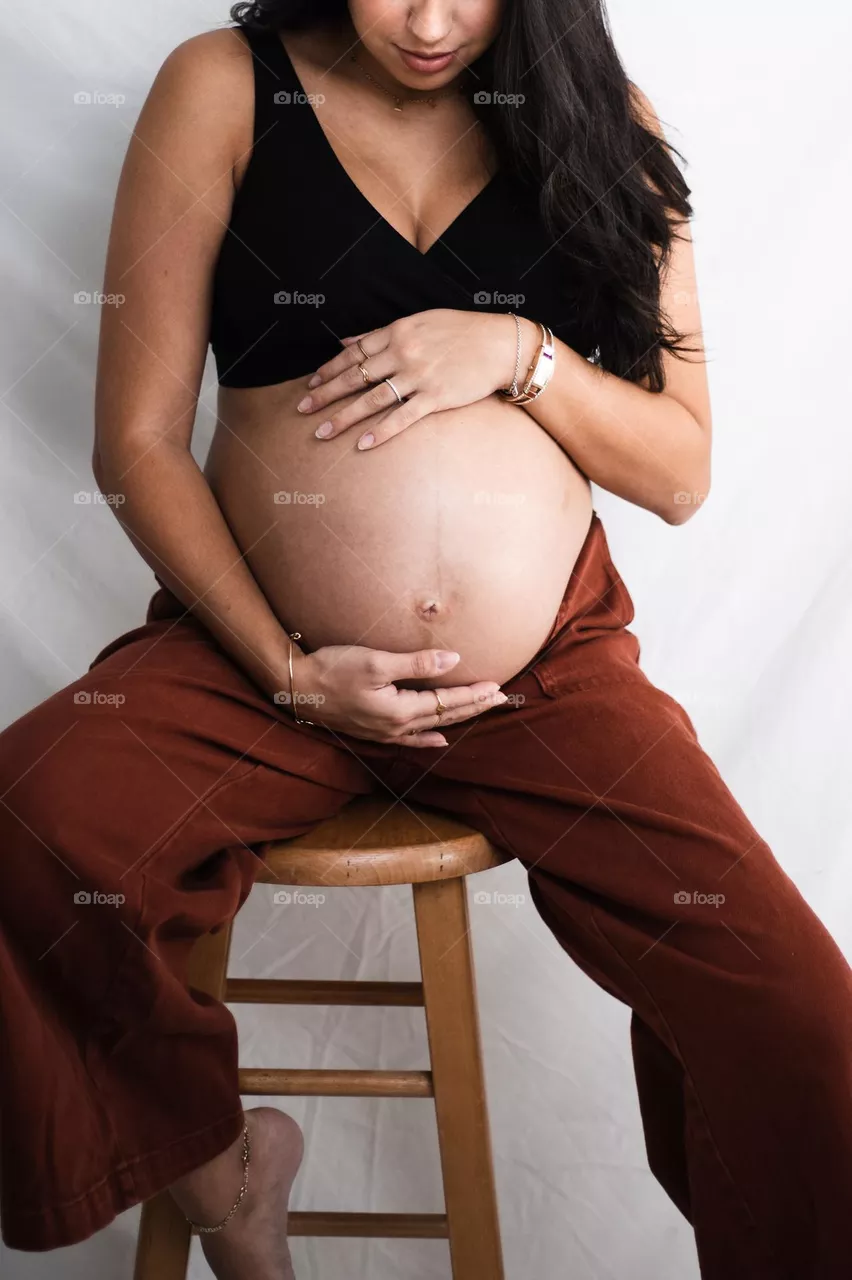 Natural beauty, a pregnant mother embracing her beautiful baby bump in front of a relaxed white backdrop as she sits on a plain wooden stool.