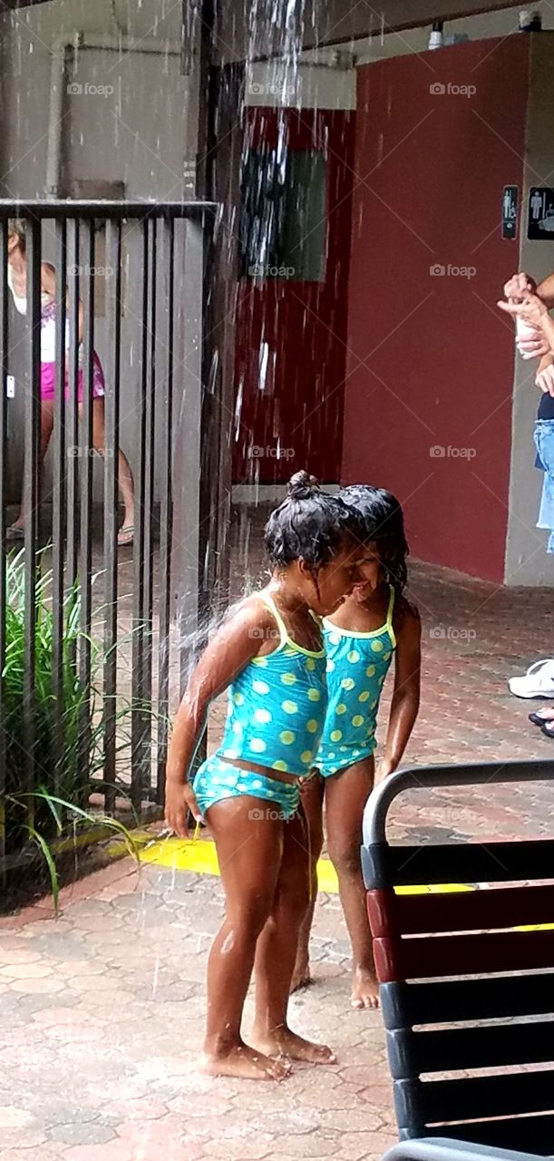 Twin girls in matching bathing suits playing in the rain together. They were giggling, splashing & standing under a rain downspout.