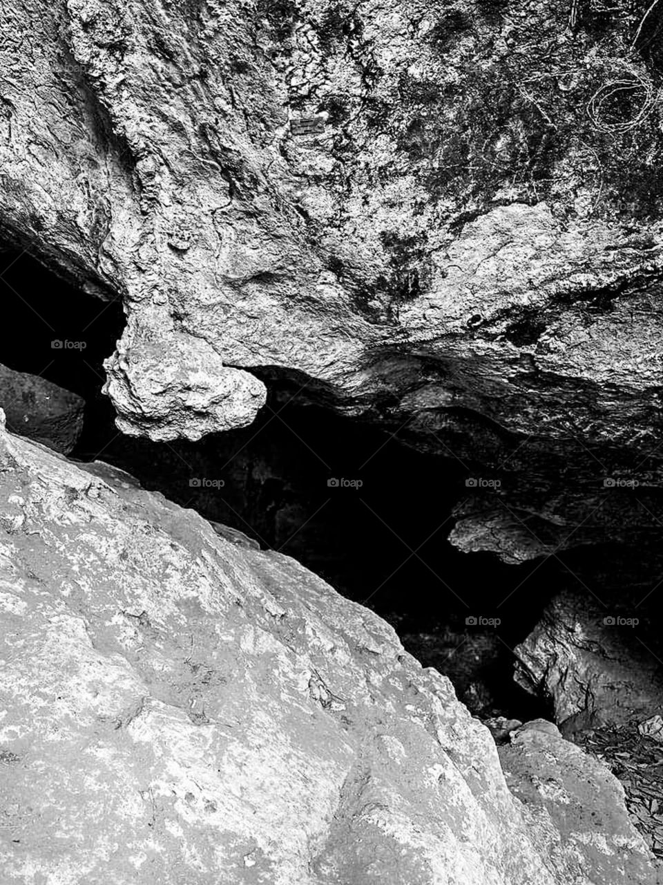 Black and white photo portrait showing the rough and hollow surface of a rock. Between the large rocks, you can see a hole or gap that leads into the cave