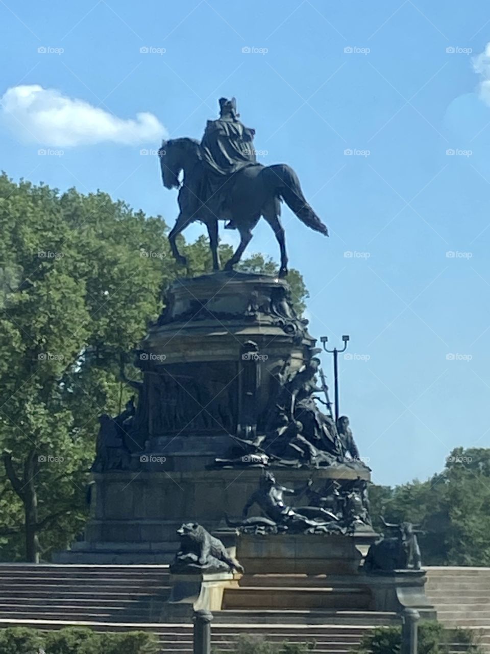 A black statue of a man on a horse against a blue sky with green trees in the background 