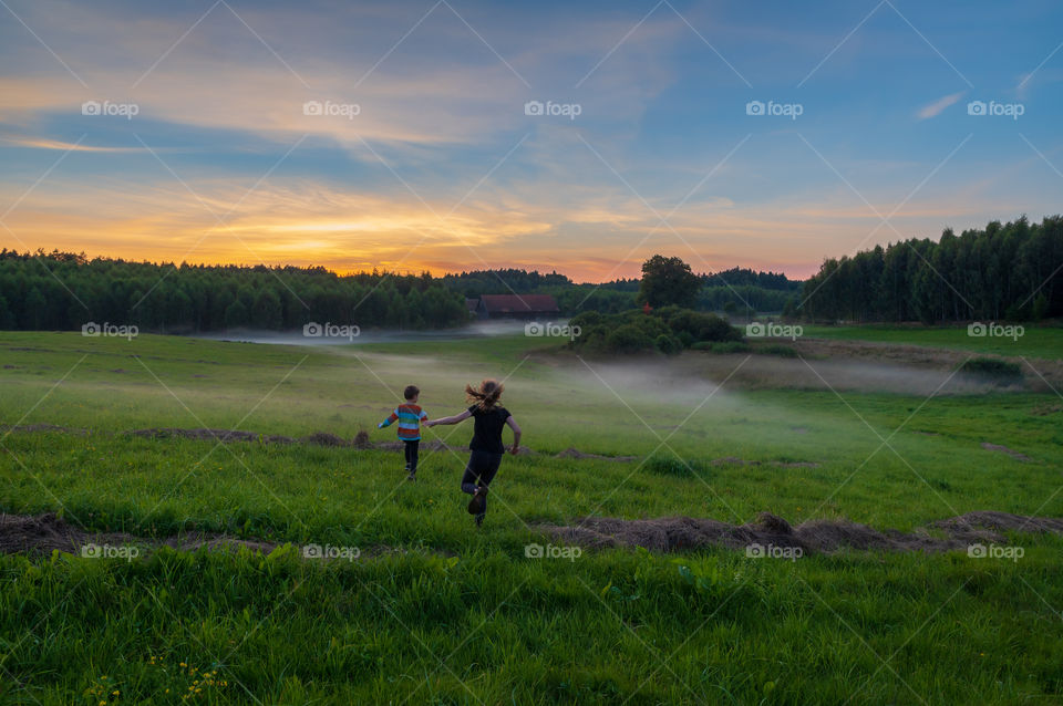 Back wiew at children running on the meadow towards ribbons of early morning fog.