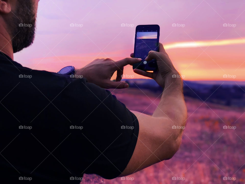 Young man taking a photo of a beautiful purple sunset over the flowering field using mobile phone 