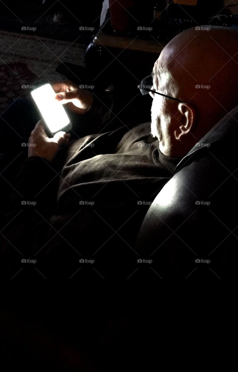Man in recliner, dark room, looking at cell phone. Monochromatic pic.