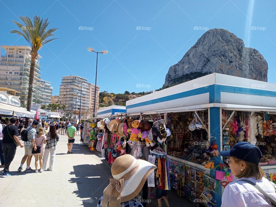 Calpe Traders Market Stalls selling Souvenirs and other items to Tourists, with Penon de Ifach otherwise known as Calpe Mountain in the background. Southern Spain.Some of the wears visible are hats and soft Toys or Dolls.