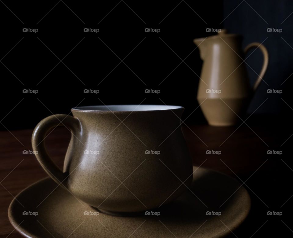 A coffee cup and saucer in a dark and shadowy setting with a coffee pot behind it