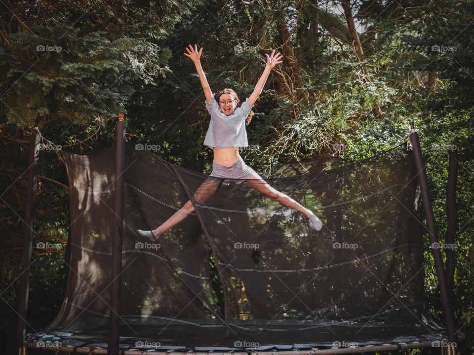 Portrait of one teenage Caucasian girl jumping and laughing on a trampoline in the back garden of a house on a summer afternoon, close-up side view. The concept is happy childhood, happy moment.