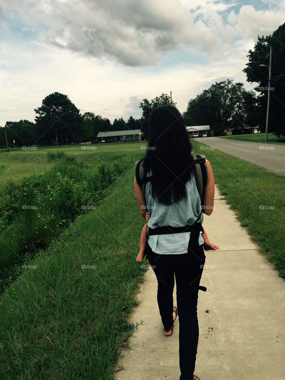 Back of young woman with long dark hair walking down the sidewalk in very small town with her baby strapped to her in front, unseen. 