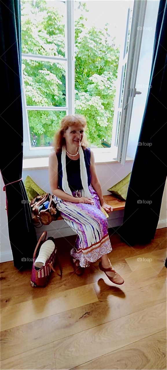 The lovely traveling lady sits inside at the window at “Schloß Blumenthal”, “Castle Blumenthal” in “Aichach”, Bavaria while she waits to be seated at the gourmet restaurant that features hearty Bavarian cuisine. 2024. Hypnotic Productions