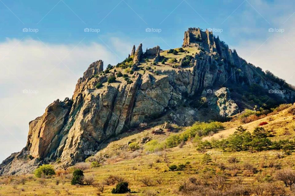 Long distance view of a towering rock mountain with green vegetation around it and blue sky in the background in low angle view