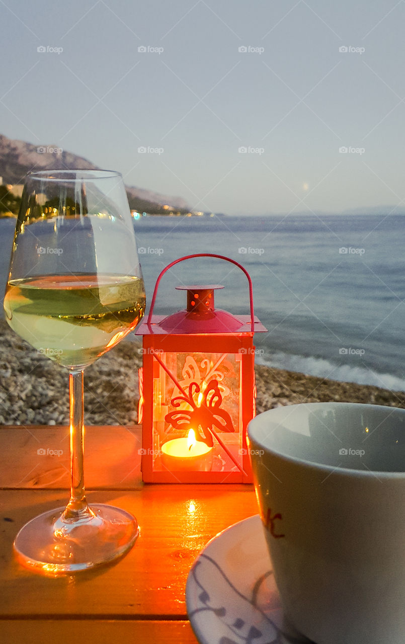 Summer evening landscape of the Dolmatian coast through a still life with a glass of white wine, a cup of tea and a candlestick with a burning candle