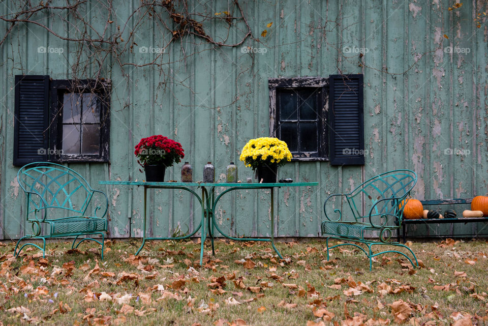 Antique rustic teal table and chairs in front of a rustic teal barn with fall leaves on the ground and mums on the table