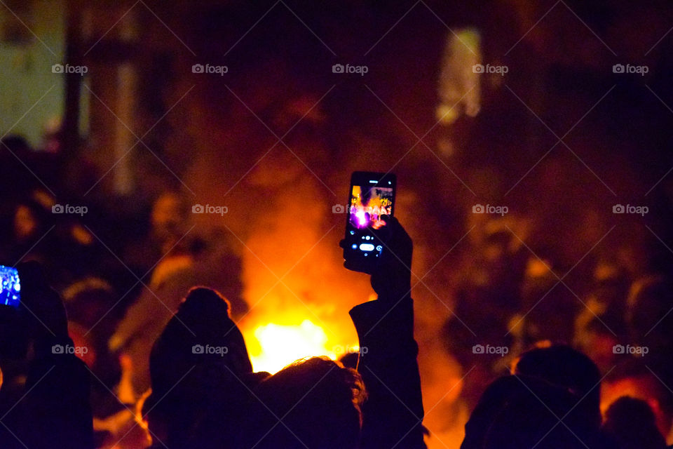 Crowd gathers around blazing tar barrel during demonstration, with on lookers filming with their cellphones.