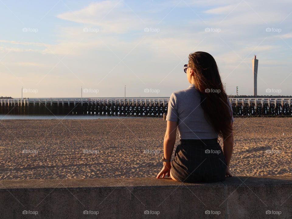 Beautiful young caucasian brunette girl with long hair sits from the back on a concrete curb admiring the sunset scenery on the north sea coast in Oostende in Belgium, close-up side view.