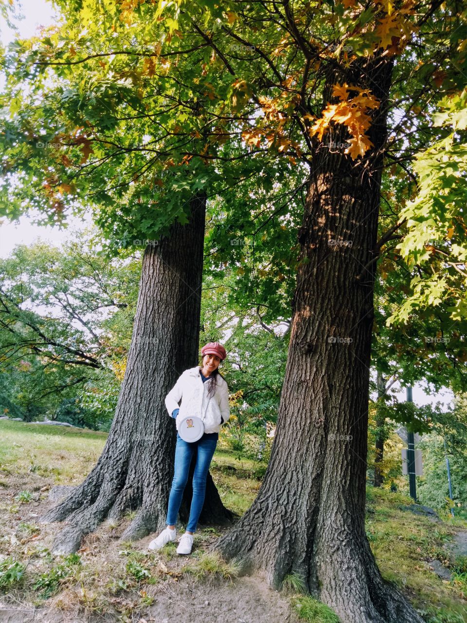 A woman wearing a red hat and white sweater, putting her hands on the pocket. Standing with two tree trunks background. 