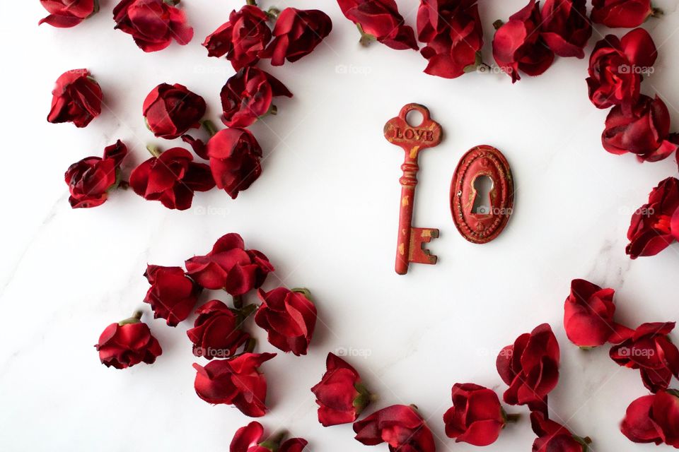 Red, metal, vintage key and keyhole with red, silk rose buds arranged in a heart-shape on white marble surface